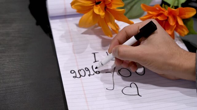 Close-up of a hand writing an I love you note with dates on lined paper, orange flowers nearby on dark desk, soft indoor lighting with shallow depth of field.
