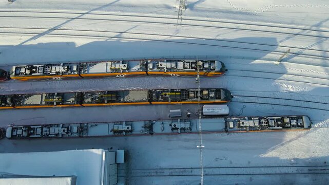 Top down aerial glide across a snow covered electric rail depot as low winter sun casts long shadows from parked rolling stock and parallel tracks during a calm sunny day.
