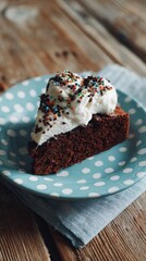 A slice of chocolate cake is placed on a blue polka-dot plate. The cake features whipped cream and colorful sprinkles on top. The setting shows a wooden table with a cloth underneath