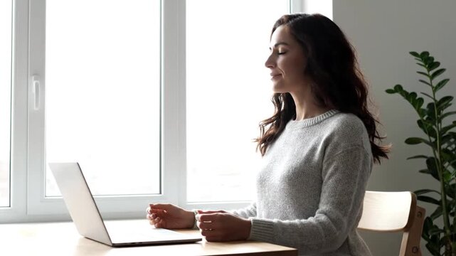 Young woman meditating or relaxing at a desk with a laptop by a bright window in a home office setting.