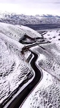 ILI, XINJIANG/CHINA - FEBRUARY 9 2026: A vertical aerial drone shot captures a black car navigating a winding asphalt highway that cuts through a pristine white snowfield. In the far distance, the maj