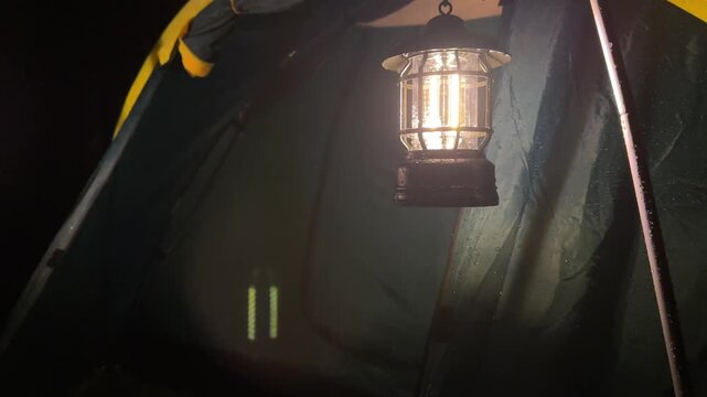 Close-up of a lit vintage camping lantern hanging inside a tent during a rainy night, symbolizing adventure, peace, and practical outdoor lighting for travel and leisure in nature.