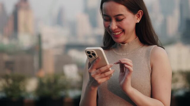 Portrait of happy Asian woman reads messages in online chat on smartphone in sunset city. Smiling Chinese female scrolls internet on mobile phone as stands in downtown block