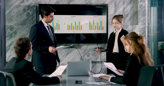 Multiracial business team in corporate meeting room discussing sales strategy with digital bar chart on screen, businesswoman presenting data while colleagues taking notes and sharing opinions