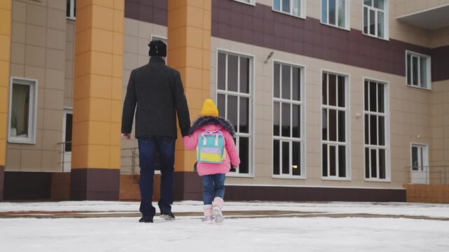 father takes child school holding his hand. happy family. snowy road school. father leads his little girl with school backpack through snow school. cold season. Dad holds hand child kid with backpack.