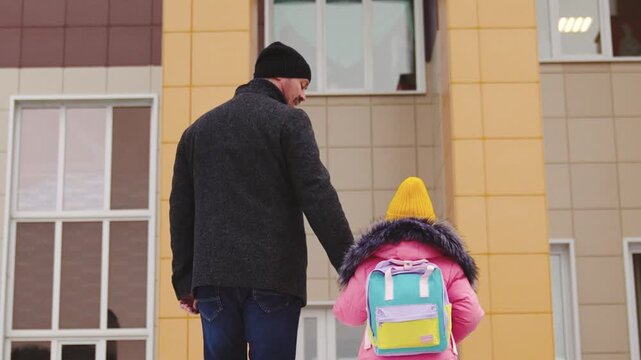 little child with backpack goes school, holding his father hand, caring young father, happy family, cheerful child girl walks with backpack into school building, snowy weather, people outdoors winter