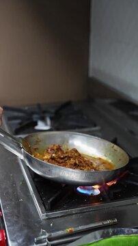 Close-up of mutton chaap frying in pan as a hand stirs over high flame.