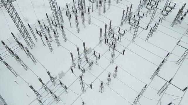 Low altitude aerial orbit above a frozen electrical substation, revealing dense rows of high voltage insulators, cables, and steel structures buried in pristine winter snow.