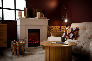 Naklejka premium Interior of living room with sofa, coffee table, lamp, fireplace and firewood in basket near burgundy wall