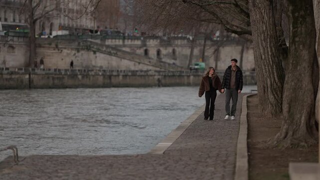 A young couple walks hand in hand along the Seine River in Paris France The romantic scene unfolds on an overcast day capturing a moment of love and connection as they stroll along the riverbank.