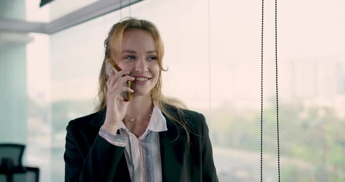 Caucasian female adult businesswoman smiling while talking on smartphone near window in office representing teamwork communication leadership in professional business planning environment