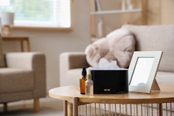 Air humidifier with bottles of essential oil and frame on coffee table near sofa in living room,...