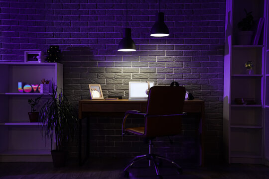 Interior of dark programmer's office with workplace, shelf units and glowing lamps at night