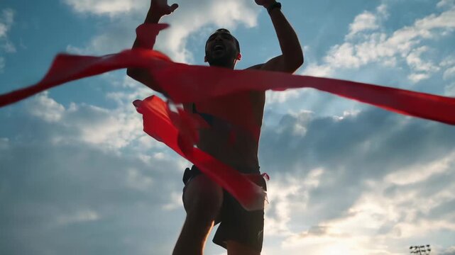Runner crosses finish line, breaking tape at track event in evening with crowd watching