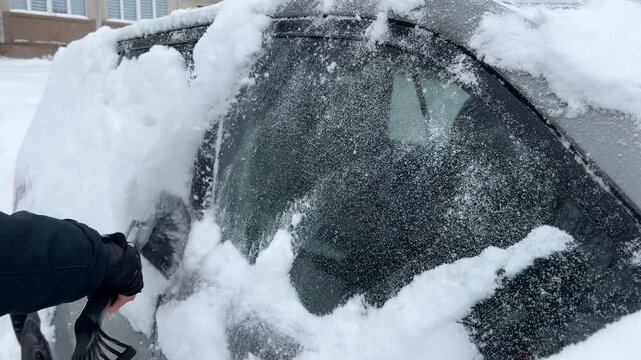 Close-up of gloved hand using snow brush to clear fresh snow from car windows. Winter morning car-preparation routine, cold-weather car maintenance, safe commuting.