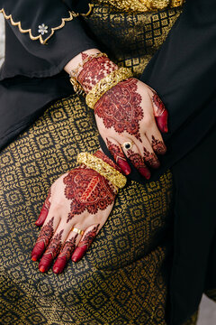 A stunning shot of a brides hands resting on a songket fabric, featuring dark henna stains, heavy gold jewelry, and traditional black and gold clothing. 