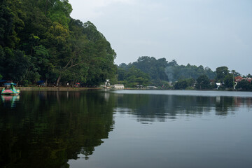 Fototapeta premium Calm lake surrounded by dense green forest and distant buildings