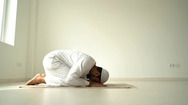 A Muslim man in traditional white Islamic attire prostrating on a prayer mat during Salat in a bright, minimalist room. Perfect video for Eid al Fitr or Ramadan.