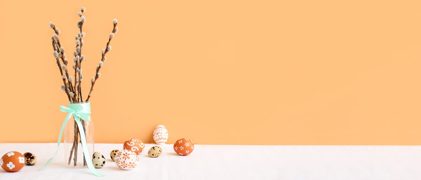 Glass vase with pussy willow branches, turquoise ribbon and Easter eggs on white table near orange wall