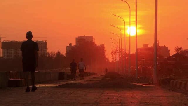 4k cinematic sunset b-roll of silhouette people walking along coast on sidewalk while sun is going down behind buildings Phan Thiet, Vietnam