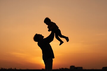 Fototapeta premium Silhouette of caucasian male adult lifting young child at sunset