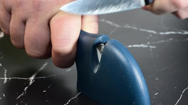 a person using a blue manual sharpener to sharpen a kitchen knife on a dark marble surface.