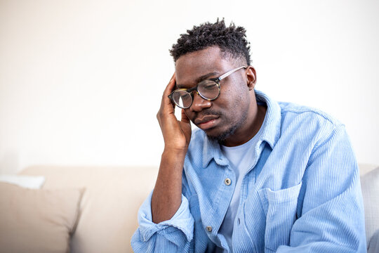 Stressed man with head pain. Black man leaning on hand with closed eyes sitting on couch. Healthcare and medicine concept. Male with migraine, fatigue, or depression. Tired person at home.