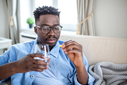 Man Taking Medicine with Water, Pill and Supplement for Treatment, Healthcare and Medicine Concept. African American man in glasses sitting on couch holding a yellow capsule and a glass of water.