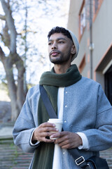 Fototapeta premium Man standing on paved walkway holding disposable coffee cup wearing grey coat green scarf bag strap