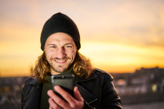 Portrait of bearded young man wearing wooly hat looking at mobile phone by sunset
