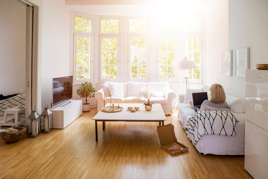 Back view of blond mature woman sitting on couch in the living room using laptop