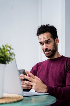Young man stting at table at home using laptop and cell phone