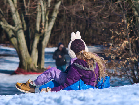 A young girl sits on a blue sledding mat, looking out over the snowy hills of Normafa, Budapest. Concept of winter relaxation, childhood joy, and active family holidays in Europe. Model released.