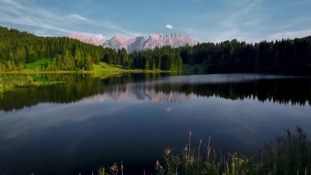 Aerial view of a serene lake mirroring distant mountains, framed by lush green forests and golden reeds in the foreground, Krun, Bavaria, Germany.
