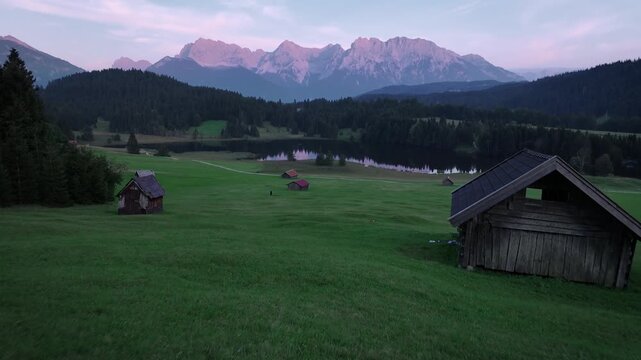 Aerial view of wooden huts scattered across a grassy field with a serene lake reflecting the sky in the distance, Krun, Bavaria, Germany.