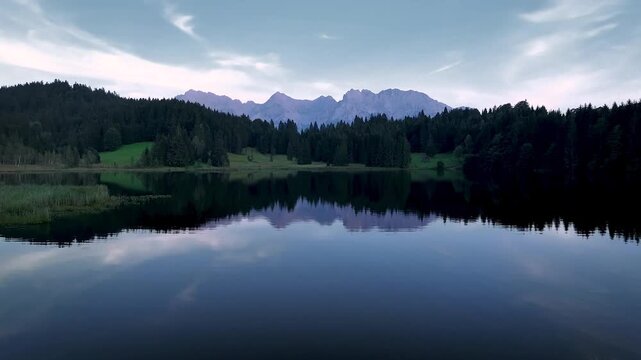Aerial view of a tranquil lake mirroring distant mountains under a dusky sky, framed by lush greenery, Krun, Bavaria, Germany.