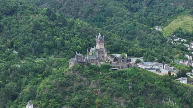 Aerial view of the striking Cochem Castle perched on a lush green hill, its grey stone contrasting beautifully with the verdant forest, Cochem, Rheinland-Pfalz, Germany.