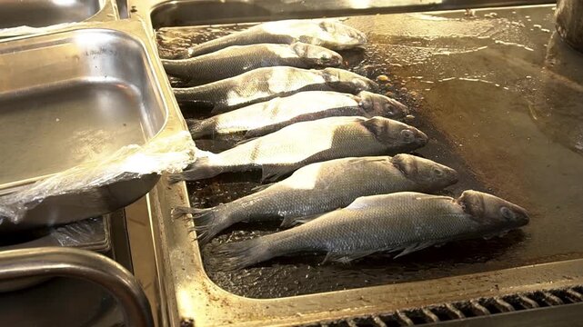 Fish is fried in a restaurant kitchen on a large baking sheet. The fish are laid out in a row. Steam rises up from the baking sheet. Perfect for content about cooking and food preparation.
