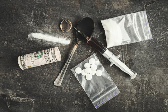 Top view, concept of drug addiction, broken life and ruined marriage, wedding ring on needle of syringe with narcotics dose, white powder, rolled banknotes, bag of pills and spoon on dark surface