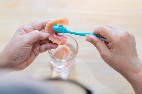 Rear view of an elderly person holds a full upper denture while cleaning it with a toothbrush