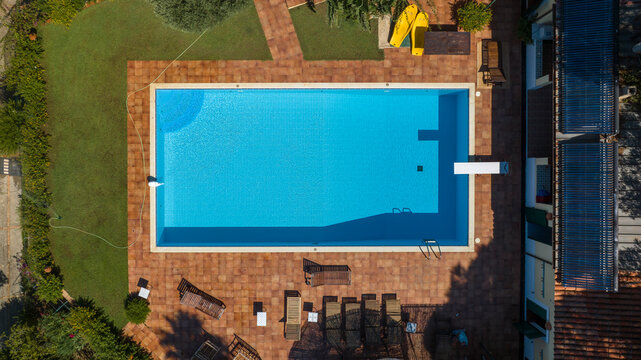 Top-down aerial view of a rectangular blue swimming pool. The pool is bordered by a terracotta-tiled patio, a diving board, and a vibrant green lawn with lounge chairs. Zenithal view of a garden.
