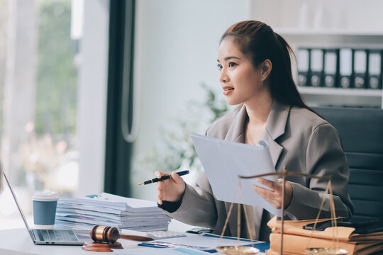 Confident Lawyer Working at Desk: A young Asian female lawyer confidently works on her laptop, surrounded by legal symbols. She exudes professionalism and competence in her modern office setting.