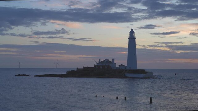 A wide cinematic shot of the historic St Mary&rsquo;s Lighthouse on its tidal island in Whitley Bay during a vibrant winter dawn. The scene features the traditional white tower and red-roofed cottages 