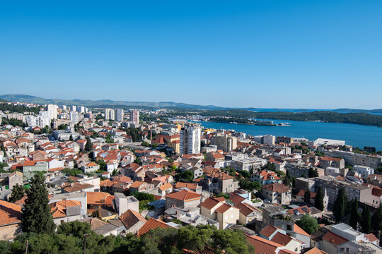 View of a vibrant cityscape where red-tiled roofs meet the azure sea under a clear blue sky, a harmonious blend of urban life and natural beauty, Sibenik, Croatia.