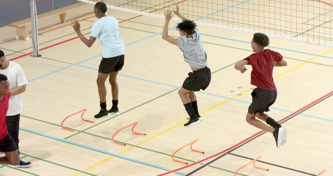 African American male coach clapping for drill, athletes jumping orange hurdles by net in gym