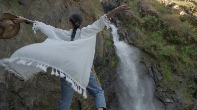 Happy Asian woman in boho dress standing on a rock with arms raised, enjoying the fresh spray of a small waterfall.