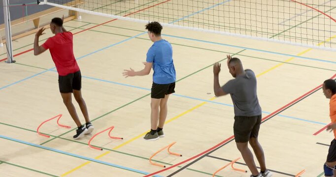 Diverse male teammates responding to coach signals, jumping over orange hurdles on gym floor
