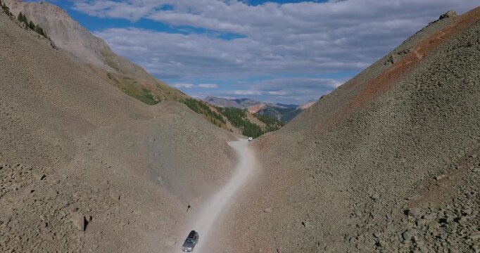Ophir Pass road threads between steep rock walls and loose debris fields, car drives down path cautiously, aerial dolly