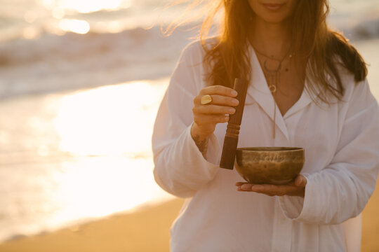 Woman playing on singing bowl standing on the beach at sunset.