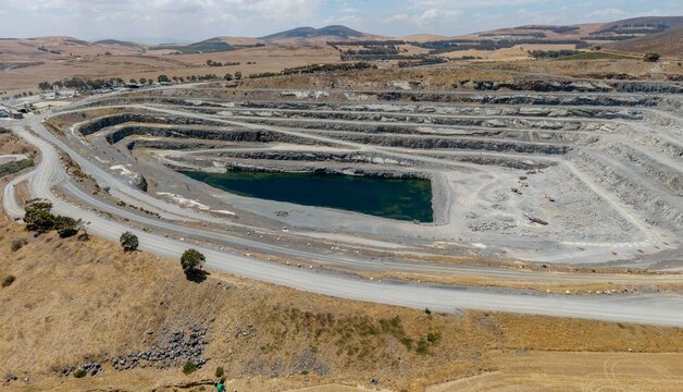 Aerial view of a vast quarry, its stepped terraces revealing layers of earth and a dark pool of water reflecting the sky, Cape Town, Western Cape, South Africa.